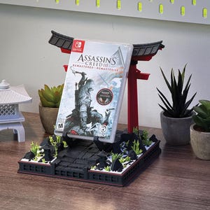 May include: A red and black decorative stand shaped like a Japanese torii gate, holding a Nintendo Switch game case. The game case features the title "Assassin's Creed III Remastered". The stand is decorated with miniature plants and rocks.