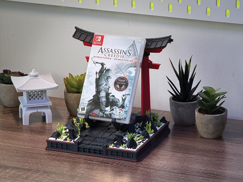 May include: A red and black decorative stand shaped like a Japanese torii gate, holding a Nintendo Switch game case. The game case features the title "Assassin's Creed III Remastered". The stand is decorated with miniature plants and rocks.