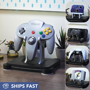 Puede incluir: Un mando gris de Nintendo 64 sobre un soporte negro, con botones rojos, verdes y amarillos. El soporte está sobre una superficie de madera. También se ven otros soportes para mandos.