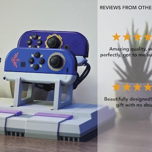 May include: A blue and white video game controller stand with a retro design. The stand holds two controllers and features a gray base with purple accents. The controllers are blue with black buttons and gold accents. The text "REVIEWS FROM OTHER STANDS" is visible.