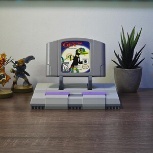 May include: A gray Nintendo 64 video game cartridge with the title "Gex" displayed on a stand resembling a Super Nintendo console. The image includes figurines and potted plants, creating a gaming-themed display.