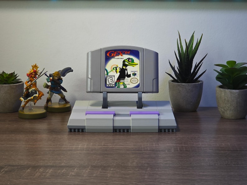 May include: A gray Nintendo 64 video game cartridge with the title "Gex" displayed on a stand resembling a Super Nintendo console. The image includes figurines and potted plants, creating a gaming-themed display.