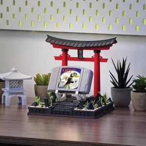 May include: A gray Nintendo 64 video game cartridge with the game title "Gex 64" displayed on a stand with a miniature Japanese Torii gate and plants. The scene is set on a wooden surface.