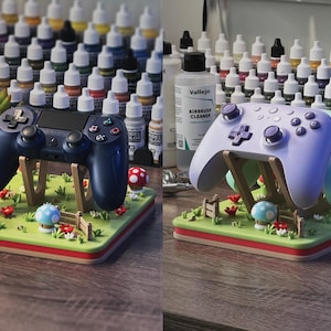 Pode incluir: Dois comandos de jogos, um azul marinho e outro lil&aacute;s claro, repousam sobre suportes decorativos. Os suportes s&atilde;o projetados como paisagens em miniatura com grama verde, flores vermelhas e detalhes de cogumelos. Frascos de tinta s&atilde;o vis&iacute;veis ao fundo.