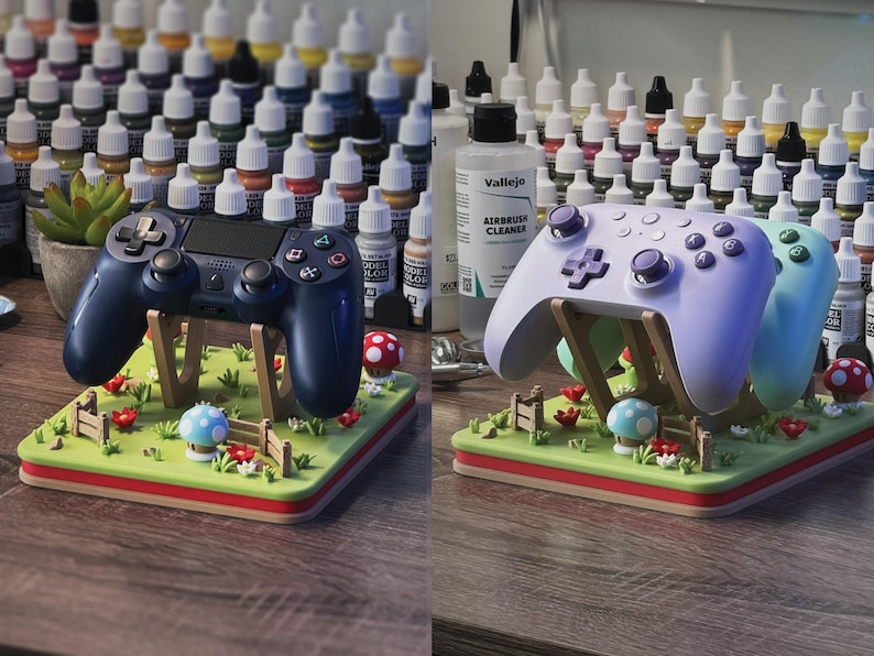 Pode incluir: Dois comandos de jogos, um azul marinho e outro lil&aacute;s claro, repousam sobre suportes decorativos. Os suportes s&atilde;o projetados como paisagens em miniatura com grama verde, flores vermelhas e detalhes de cogumelos. Frascos de tinta s&atilde;o vis&iacute;veis ao fundo.