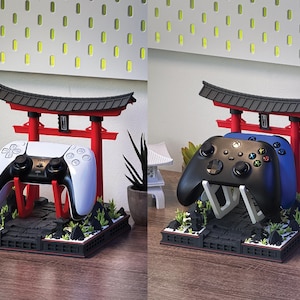 May include: A gaming controller stand featuring a red and black Japanese torii gate design. The stand holds a white and black PlayStation 5 controller. The base is black with green and gray accents, and the stand is on a wooden surface.