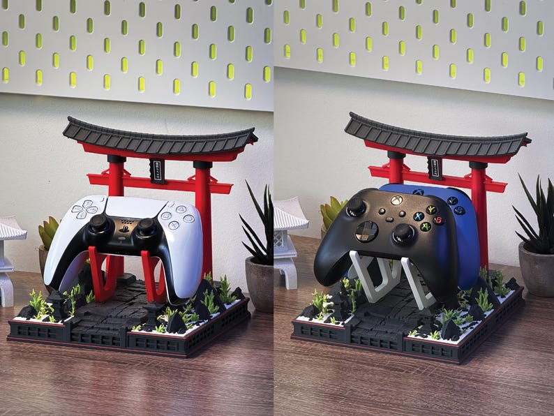 May include: A gaming controller stand featuring a red and black Japanese torii gate design. The stand holds a white and black PlayStation 5 controller. The base is black with green and gray accents, and the stand is on a wooden surface.