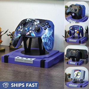 May include: A purple video game controller stand with a controller featuring a blue and white design. The stand is shaped like a classic game console. Additional controller stands are shown in the image.