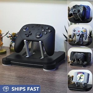 May include: Black gaming controller stand with a black controller on top. The stand is designed to resemble a classic game console. The image also shows other game controllers and a game cartridge.
