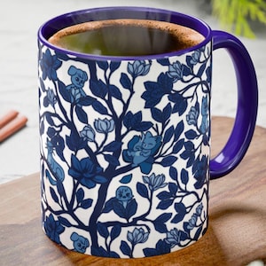 Pu&ograve; includere: Una tazza di ceramica bianca con un motivo floreale blu e un manico e interno viola. La tazza &egrave; riempita con un liquido scuro, forse caff&egrave; o t&egrave;, ed &egrave; appoggiata su un tagliere di legno.