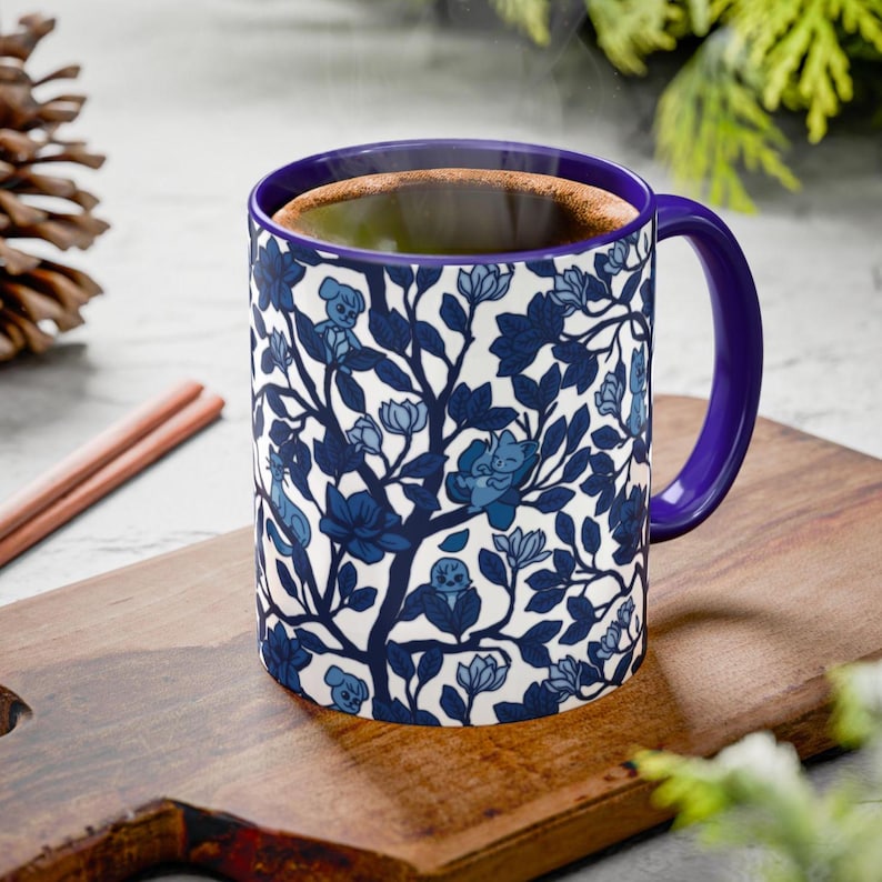 Pu&ograve; includere: Una tazza di ceramica bianca con un motivo floreale blu e un manico e interno viola. La tazza &egrave; riempita con un liquido scuro, forse caff&egrave; o t&egrave;, ed &egrave; appoggiata su un tagliere di legno.