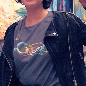 Op de afbeelding: Een vrouw draagt een grijs T-shirt met een regenboogkleurige afbeelding met de tekst "Cinema" en een zwart leren jack.