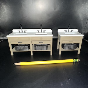 以下が含まれることがあります： 白い洗面台、タン色のキャビネット、黒い籐のバスケットを備えたミニチュアのバスルーム化粧台。化粧台には銀色の蛇口があり、暗い背景に配置されています。黄色の鉛筆が前景にあります。