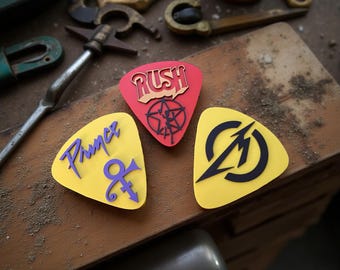 May include: Three guitar picks with band logos: "RUSH" in red, a yellow pick with "Prince" and a symbol, and a yellow pick with the "Metallica" logo. The picks are on a wooden surface with metal tools in the background.