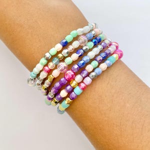 May include: A stack of colourful beaded bracelets in shades of pink, purple, blue, green, and white. The bracelets are made of small, round beads and are layered on top of each other.