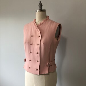 May include: A pink sleeveless vest with a double-breasted button closure and a ruffled collar.