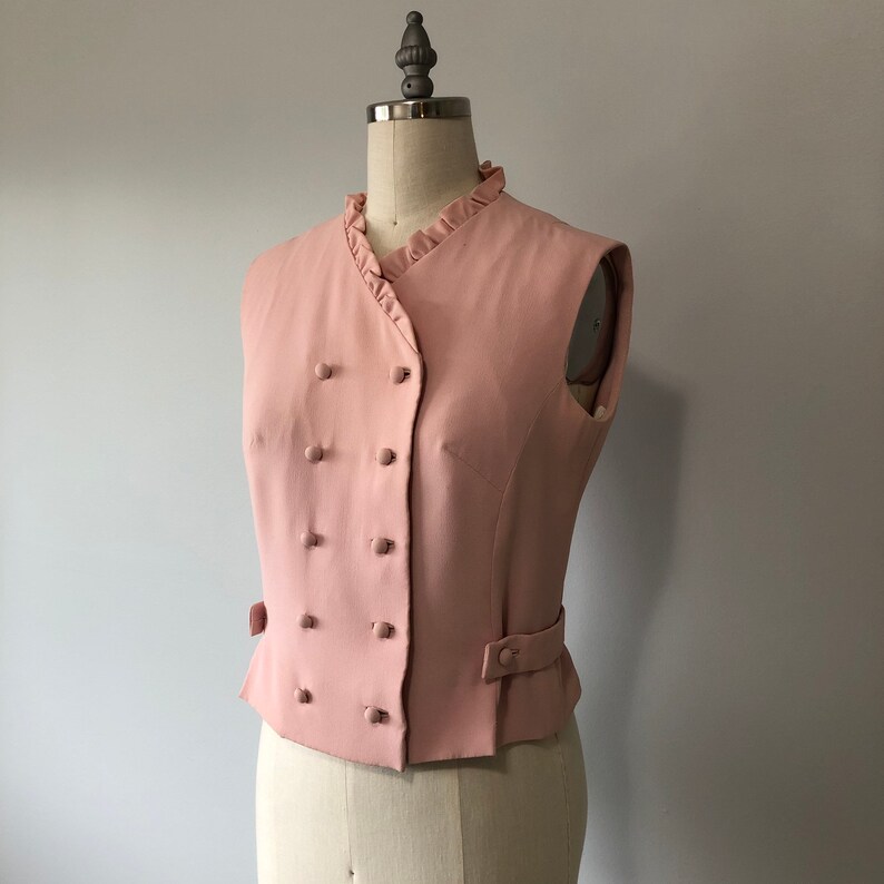 May include: A pink sleeveless vest with a double-breasted button closure and a ruffled collar.