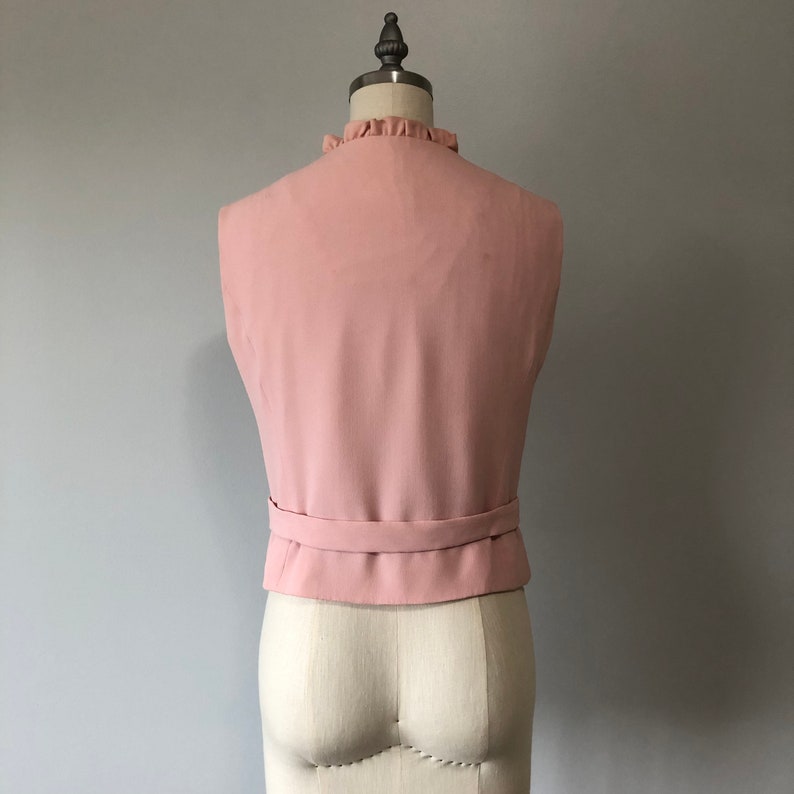 May include: A sleeveless, blush pink vest with a ruffled neckline and a matching belt. The vest is on a white dress form.