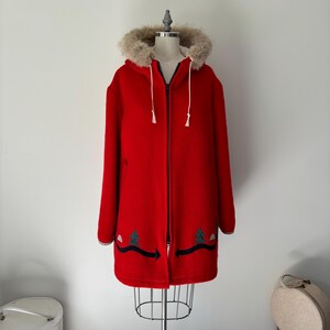 May include: A red hooded winter coat with a faux fur trim around the hood. The coat has a zipper closure and is decorated with embroidered green trees and a black arrow on the bottom.