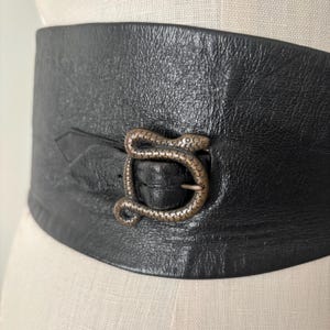 May include: Wide black leather belt with a textured surface. The belt features a silver-coloured buckle in the shape of a snake. The belt is designed to be worn around the waist. The belt is a fashion accessory.