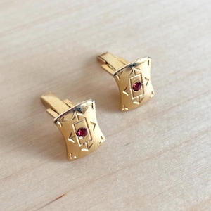 May include: A pair of gold-toned cufflinks with a rectangular design. Each cufflink features a central, round, red gemstone and an etched pattern. The cufflinks are displayed on a light-colored wooden surface.