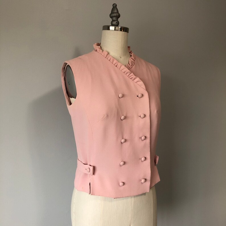 May include: A sleeveless pink vest with a double breasted button closure and a ruffled collar.