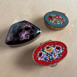 May include: Three decorative trinket boxes. One is black with pink floral designs, another is oval-shaped with a mosaic of red, white, and blue flowers, and the third is oval-shaped with a mosaic of flowers on a turquoise background.