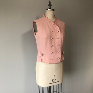 May include: A pink sleeveless vest with a ruffled collar and a double-breasted button closure.