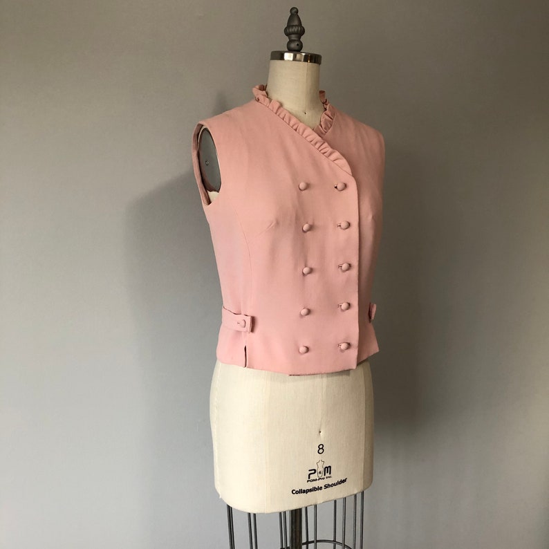 May include: A pink sleeveless vest with a ruffled collar and a double-breasted button closure.