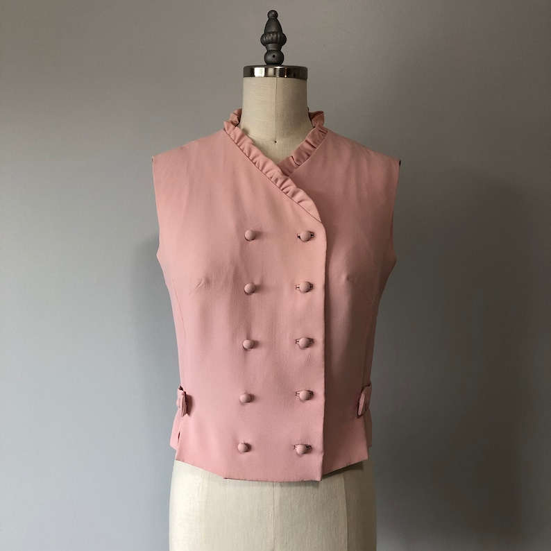 May include: A pink sleeveless vest with a double-breasted button closure and a ruffled neckline.