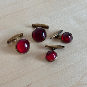 May include: Four vintage cufflinks with a gold-tone metal finish and vibrant red cabochon stones. The cufflinks are displayed on a light wooden surface, showcasing their classic design and rich colour.