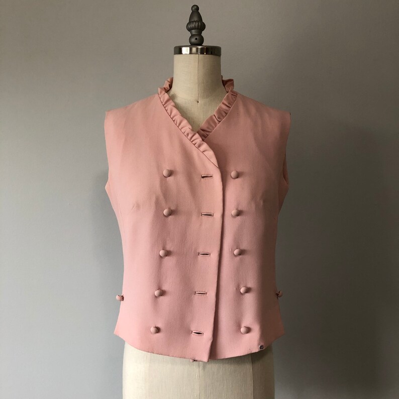 May include: A sleeveless pink vest with a ruffled collar and a double row of buttons down the front.