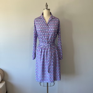 May include: A purple dress with a white and pink geometric pattern. The dress has a collared neckline, long sleeves, and a belt.