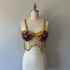 May include: A purple and gold sequined bra top with fringe. The bra top has a scalloped bottom edge and is made of sequins and beads.