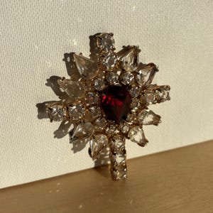 May include: A gold-toned brooch in a starburst design, featuring a large, teardrop-shaped, deep red gemstone at the center. Clear, faceted crystals surround the red stone and extend outward, creating a sparkling, elegant accessory.