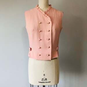 May include: A sleeveless pink vest with a ruffled collar and a double-breasted button closure. The vest has a slightly fitted silhouette and is made of a smooth fabric.