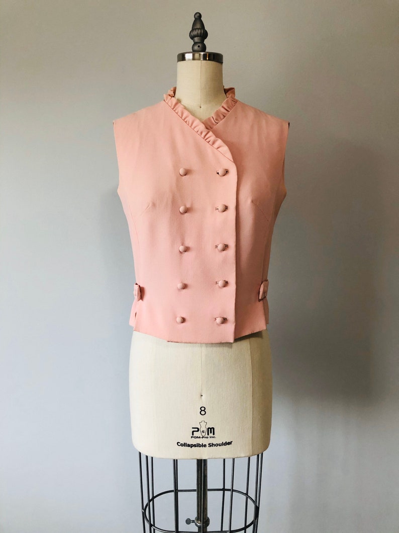 May include: A sleeveless pink vest with a ruffled collar and a double-breasted button closure. The vest has a slightly fitted silhouette and is made of a smooth fabric.