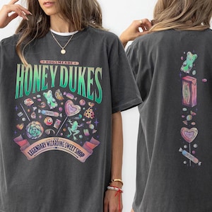 以下が含まれることがあります： カラフルなお菓子と「HONEY DUKES LEGENDARY WIZARDING SWEET SHOP」の文字がプリントされた濃いグレーのTシャツ。Tシャツの背面には、お菓子の縦型デザインが施されています。このシャツはオーバーサイズです。