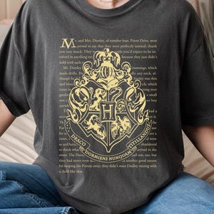 May include: Dark grey t-shirt featuring the Hogwarts crest in gold with text from the Harry Potter series. The shirt has a relaxed fit and is paired with blue jeans. The word "Pepper" is at the bottom left.