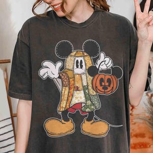 以下が含まれることがあります： パッチワークデザインのミッキーマウスのグラフィックが特徴の濃いグレーのTシャツ。カボチャ型のバッグを持っています。漫画のキャラクターは大きな黄色の靴と白い手袋をしています。シャツはヴィンテージで色あせた外観です。