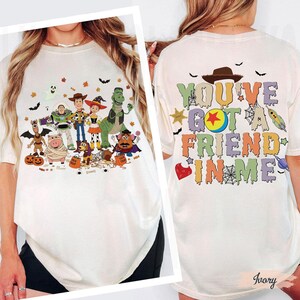 Puede incluir: Camiseta de color marfil con un gráfico colorido de personajes de Toy Story con disfraces de Halloween. La parte posterior de la camiseta tiene el texto "YOU'VE GOT A FRIEND IN ME" en varios colores con acentos temáticos de Halloween.