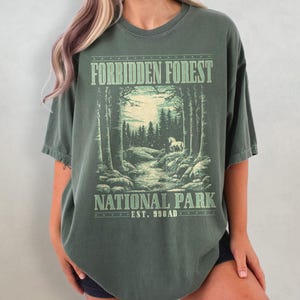 Könnte beinhalten: Olivgrünes T-Shirt mit einem Waldmotiv. Der Aufdruck enthält den Text "Forbidden Forest National Park Est. 990 AD" im Vintage-Stil. Das Design zeigt Bäume, einen Fluss und ein weißes Pferd.