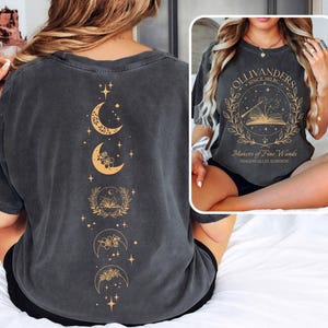 Pode incluir: T-shirt cinzenta escura com um design dourado de lua crescente e estrelas nas costas. A frente da camisa tem um gráfico circular com o texto "OLLIVANDERS SINCE 382 BC" e "Makers of Fine Wands, Diagon Alley, London."