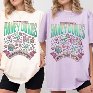 May include: Ivory and orchid-colored t-shirts featuring a colorful graphic design. The design includes the text "Honeydukes" above a collection of candies and sweets, with a banner that reads "Legendary Wizarding Sweet Shop."