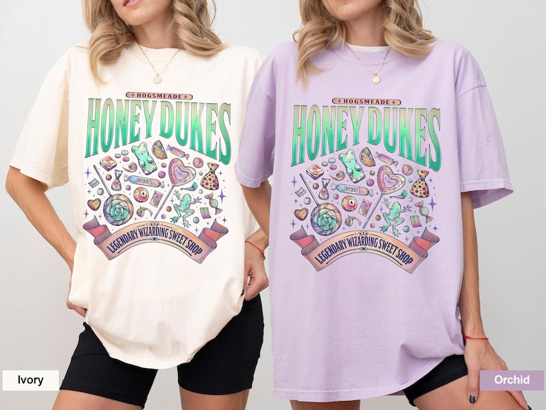 May include: Ivory and orchid-colored t-shirts featuring a colorful graphic design. The design includes the text "Honeydukes" above a collection of candies and sweets, with a banner that reads "Legendary Wizarding Sweet Shop."