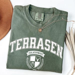 May include: A sage green short-sleeve t-shirt featuring the white text TERRASEN in an arched font above a shield emblem with the text GALATHYNIUS. The shield displays a stag and a heart. A thin gold chain necklace rests on the collar area.