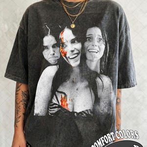 May include: A charcoal gray acid-wash t-shirt featuring a black and white graphic print of three faces with varying expressions, including one with red blood-like accents on the face and chest. The shirt has short sleeves and a crew neckline.