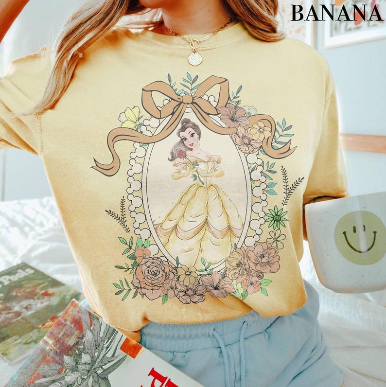 Może przedstawiać: Jasnoż&oacute;łta koszulka z oprawioną ilustracją księżniczki w ż&oacute;łtej sukni, otoczonej kwiatami i kokardą. Słowo "BANANA" jest wydrukowane czarnym kolorem w prawym g&oacute;rnym rogu. Koszulka jest noszona z niebieskimi szortami.