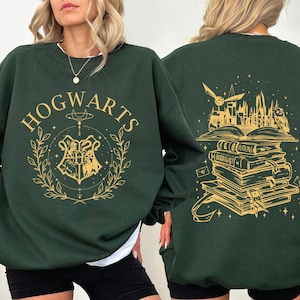 Könnte beinhalten: Dunkelgrünes Sweatshirt mit goldfarbenem "HOGWARTS"-Schriftzug auf der Vorderseite und einem Wappen-Design. Auf der Rückseite befindet sich eine goldfarbene Illustration von Büchern, Hogwarts-Schloss und einer Eule. Das Sweatshirt hat lange Ärmel und einen Rundhalsausschnitt.