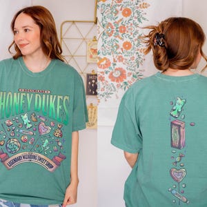 May include: A teal t-shirt with a graphic of sweets and the text "Honeydukes Legendary Wizarding Sweet Shop." The back of the shirt features a gummy bear, candy box, and other sweets. The shirt is a comfortable, oversized fit.